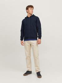 Marineblå hettegenser med glidelås, frontlomme og snor. Brukt med beige bukser og mørke joggesko, på en nøytral bakgrunn.