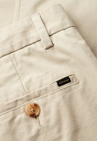 Beżowe bawełniane spodnie mają gładką fakturę, wyraźne zapięcie na guzik oraz czarną metkę z napisem "SUPERDRY" złotymi literami na tylnej kieszeni.