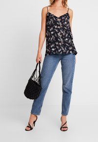 Camisole à boutons à motifs floraux noirs avec fines bretelles, associée à un jean bleu droit ; le modèle tient un sac tissé noir texturé et porte des talons noirs.