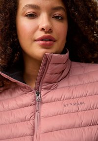 Lyserød pufferjakke med høj krave, med vandrette quiltede sektioner og sort indvendig foret. Lynlås foran og brandlogo synligt.