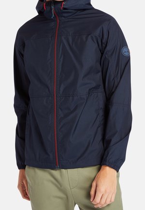 Homme portant une veste à capuche bleu marine avec une fermeture éclair rouge et un pantalon kaki, debout les mains détendues le long du corps.