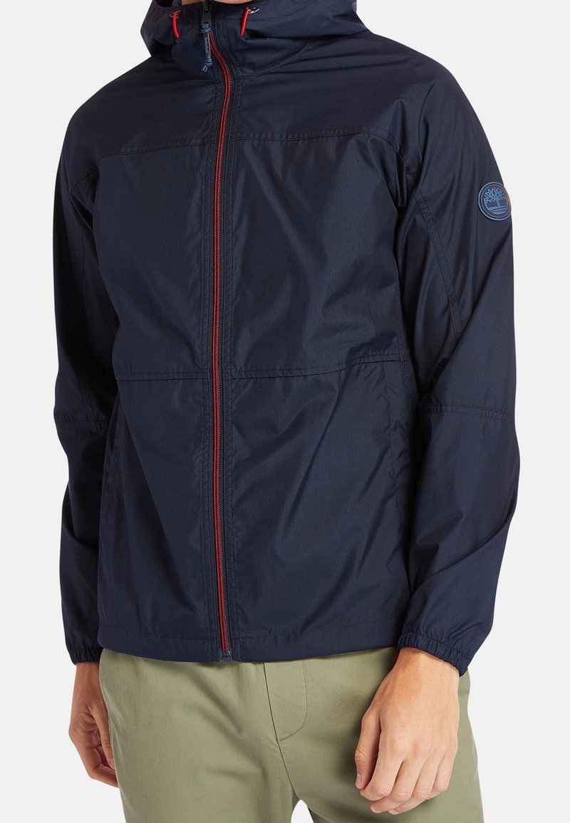 Homme portant une veste à capuche bleu marine avec une fermeture éclair rouge et un pantalon kaki, debout les mains détendues le long du corps.