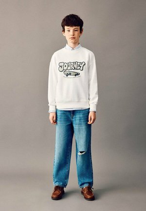 Hanorac alb cu imprimeu „JOURNEY” și skateboard, asortat cu o cămașă deschisă la culoare cu guler, blugi albaștri cu aspect uzat și pantofi maro.