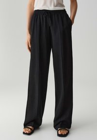 Zwarte, lichte, losse broek met een trekkoord in de taille, rechte broekspijpen en zijzakken. Soepele textuur met een subtiele glans.