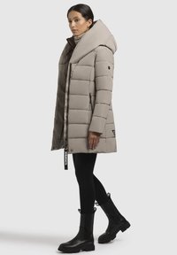 Manteau en duvet beige avec un col montant et une grande capuche, design matelassé, quincaillerie noire, poches latérales et étiquette de logo. Porté avec des leggings noirs et des bottes.