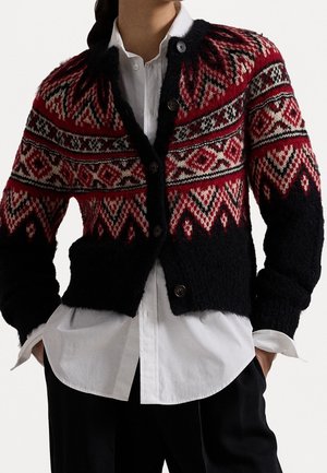 Cardigan tricoté à motifs noirs, rouges et blancs porté sur une chemise blanche boutonnée, mains dans les poches de pantalons noirs.