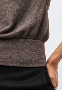 Pruun kootud top, millel on elastne vöökoht, kantud mustade pükste kohal, käsivars puusale toetumas ja külje peal nähtav.