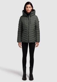 Grüne Pufferjacke mit abnehmbarer Kapuze, Chevron-Steppung, Frontreißverschluss und Seitentaschen, getragen über schwarzen Leggings und Stiefeln.