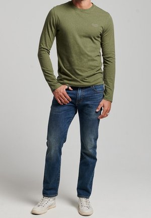 Camisa de manga larga de color oliva con cuello redondo, combinada con vaqueros azules y zapatillas blancas. La tela parece suave con una ligera textura.