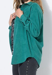 Chemise en velours côtelé teal avec un devant boutonné, des manches longues et un ourlet incurvé. Des boutons en argent notables décorent les poignets et les côtés.