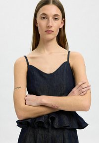 Denim peplum top s výstřihem ve tvaru srdce, tenkými ramínky a volánkovým lemem. Tmavě indigo barva, hladká textura s viditelnými švy.