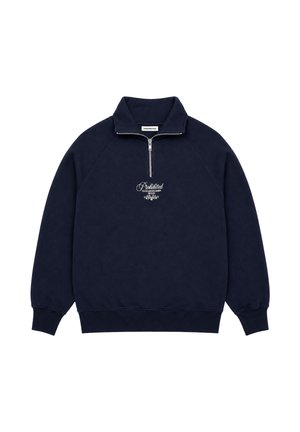 Junger Mann mit Ohrringen, der einen marineblauen Quarter-Zip-Sweatshirt über einem blau-weiß gestreiften Hemd vor einem schlichten grauen Hintergrund trägt.