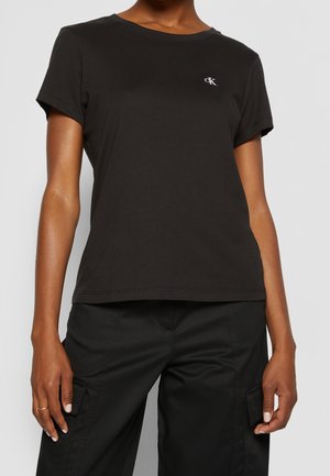Persoon draagt een zwart T-shirt met korte mouwen met een klein wit CK-logo op de borst en zwarte cargobroek met zakken.