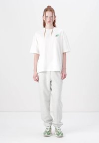 T-shirt blanche oversized avec un logo vert, associée à un sweat-pantalon gris. Le modèle porte des baskets vertes et blanches. Design minimaliste, texture lisse.