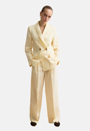 Femme portant un blazer croisé crème et un pantalon large assorti avec des chaussures noires, debout les mains dans les poches.