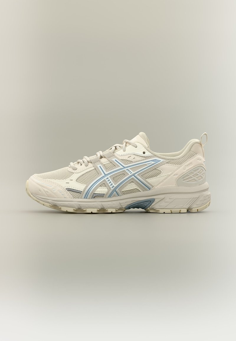 Zapatilla deportiva beige con parte superior de malla y sintética, detalles de rayas azul claro, suela crema y cierre con cordones, mostrada desde un lateral sobre un fondo neutro.