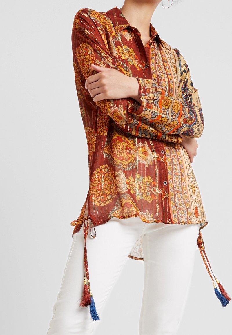 Personne portant un chemisier à manches longues à motifs rouges, oranges et beiges, associé à un pantalon blanc et des pampilles pendantes à l'ourlet.
