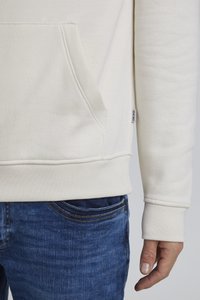 Lys beige sweatshirt med en lomme foran, ribbede manchetter og kant; parret med blå denimjeans. Materialet ser blødt og struktureret ud.