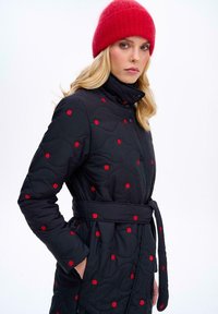 Prešívaný čierny kabát s červenými vzormi čerešní, vysokým golierom a opaskom na zaviazanie. Obsahuje štruktúrovaný povrch a bočné vrecká.