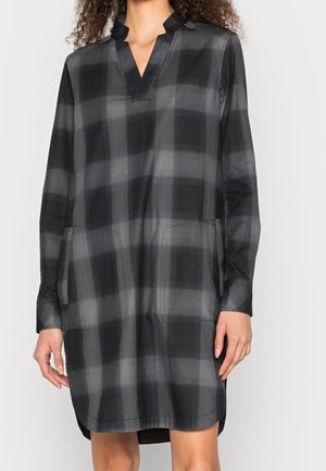 Frau trägt ein langärmeliges schwarz-graues kariertes Kleid mit V-Ausschnitt und Stehkragen und steht vor einem schlichten Hintergrund.