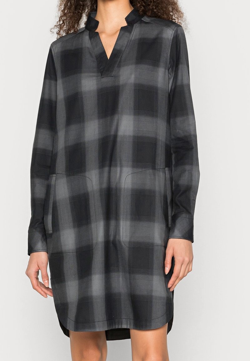 Femme portant une robe à carreaux noire et grise à manches longues avec un col en V et un col montant, debout devant un fond uni.