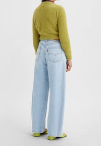 Grön fluffig tröja, hög midja på ljusblå vida jeans, synlig sömnad, två bakfickor med röd logotab, gröna klacksandaler med blockklack.