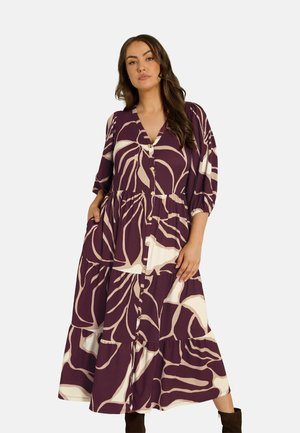 Mujer que lleva un vestido largo morado con grandes motivos de hojas beige, con botones en la parte delantera, mangas tres cuartos y bolsillos laterales, de pie con las manos en los bolsillos.