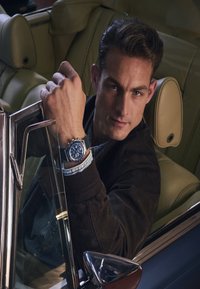 Man in dark jacket sitting in a convertible car, resting his arm on the door, wearing a blue chronograph wristwatch.
