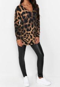 Femme portant une tunique à manches longues à motif léopard marron et noir, des leggings en cuir noir et des baskets blanches épaisses, debout devant un fond blanc.