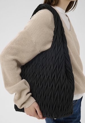 Persona, kas valkā bēša krāsas adītu džemperi un zilas džinsas, tur lielu melnu teksturētu plecu somu uz labā pleca.