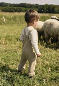 Salopette en tricot beige avec des bretelles, associée à une chemise blanche à manches longues et des poignets retroussés, portée par un enfant dans un champ herbeux.