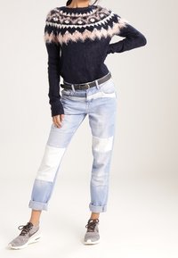 Person som bär en fluffig marinblå tröja med mönster på oket, ljusa patchwork-jeans uppvikta vid anklarna, ett svart bälte och grå sneakers med snörning.