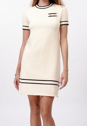 Femme portant une robe crème à manches courtes avec des bordures noires aux manches, au col, à l'ourlet et une petite poche poitrine avec une broche.