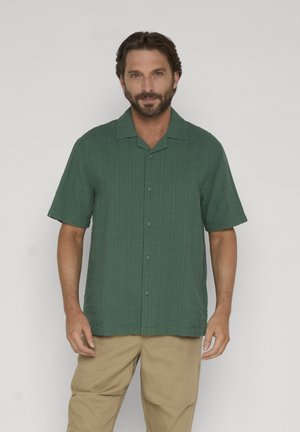 Homme aux cheveux courts et foncés avec une barbe, portant une chemise verte à manches courtes boutonnée et un pantalon beige, se tenant devant un fond uni.