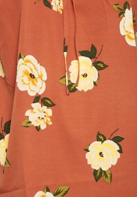 Tkanina w kolorze rdzy z białymi i żółtymi wzorami kwiatowymi oraz zielonymi liśćmi i detalem zawiązywanym na szyi. Gładka tekstura i casualowy design.