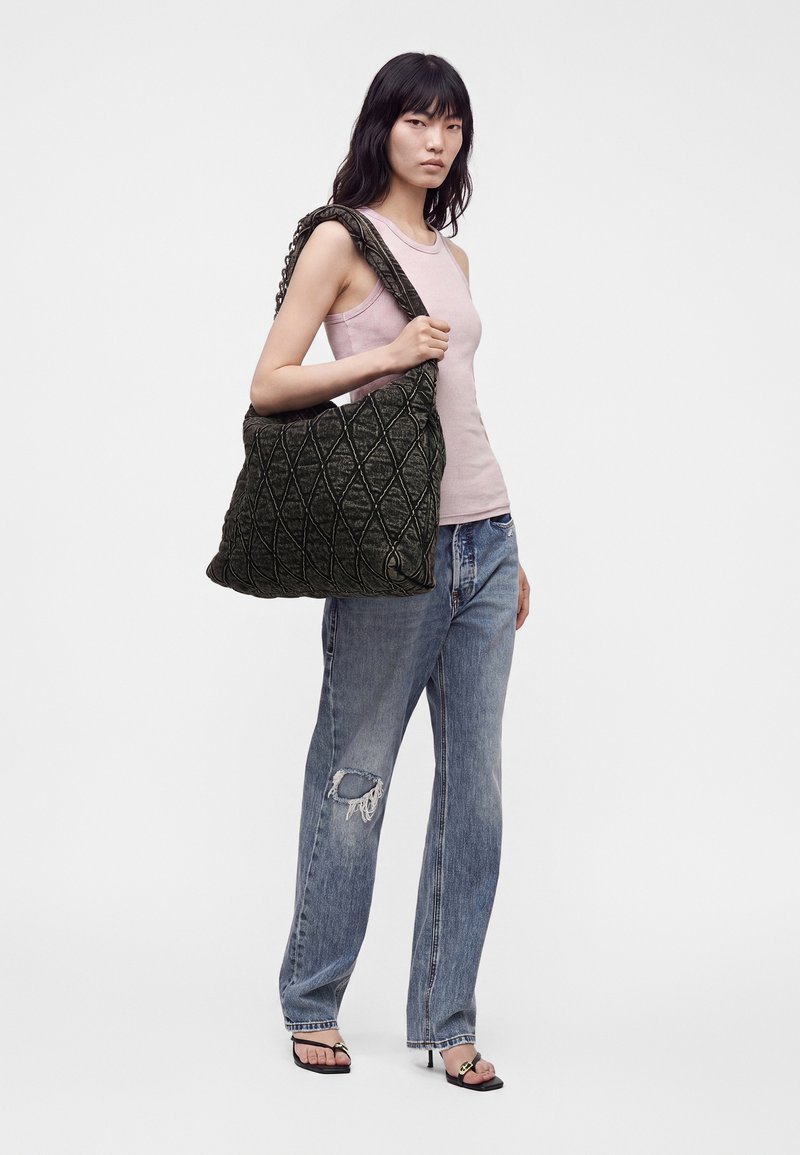 Femme portant un haut rose sans manches et un jean bleu déchiré tient un grand sac à épaule noir matelassé, debout devant un fond blanc uni.
