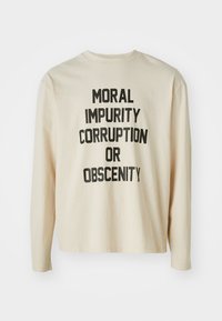 Beige langermet skjorte laget av bomull, med fet svart tekst som leser "MORALSK URENSKAP KORRUPSJON ELLER OBSCENITET" i varierende størrelser.