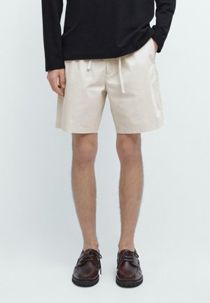 Person trägt beige Shorts mit Kordelzug, schwarzen Langarmtop und dunkelbraune Lederschnürschuhe, steht mit einer Hand in der Tasche.