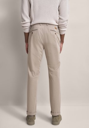 Person trägt beigefarbene Chinos mit geknöpften Gesäßtaschen, beigefarbenen Pullover mit hochgekrempelten Ärmeln und beigefarbene Turnschuhe, steht vor neutralem Hintergrund.