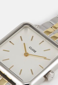 Montre en métal carrée avec un bracelet en argent et or, cadran blanc, index heures en or et aiguille des secondes en or. Nom de la marque "CLUSE" affiché.