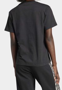 Person som bär en enkel svart kortärmad t-shirt och svarta byxor med leopardmönster och randig sidodetalj, visad bakifrån.