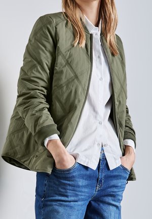 Osoba ubrana w zieloną pikowaną kurtkę na suwak, noszoną na białej koszuli zapinanej na guziki, z rękami w kieszeniach niebieskich dżinsów, stojąca na jednolitym tle.