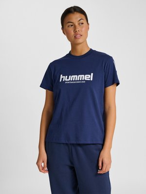 Vrouw draagt marineblauw Hummel T-shirt en bijpassende broek, staat tegen een effen lichte achtergrond en kijkt opzij.