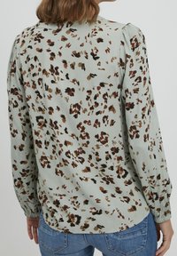 Blouse à manches longues avec une base vert clair, ornée d'un imprimé animalier marron et noir. Détail plissé aux épaules et poignets élastiques.
