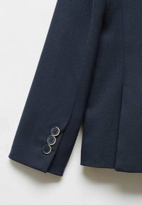 Manica di blazer blu navy con tessuto strutturato, caratterizzata da tre bottoni neri con dettagli dorati. Si notano il taglio sartoriale e le linee pulite.