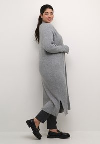 Lunga cardigan grigia a costine con fessure laterali, indossato sopra pantaloni scuri aderenti e mocassini neri con dettagli in metallo dorato.