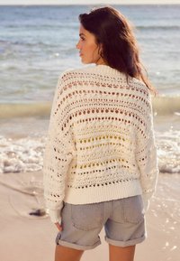 Un pull en tricot blanc avec un motif ajouré, doté d'un col haut et de manches longues, associé à un short en denim clair. L'océan en arrière-plan.