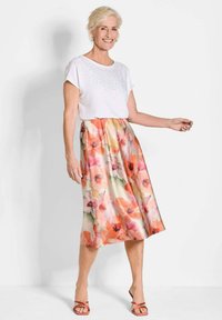 Weißes T-Shirt mit kurzen Ärmeln und Strassverzierungen, kombiniert mit einem floral bedruckten Rock in Orange- und Rosatönen, ergänzt durch rote Sandalen.