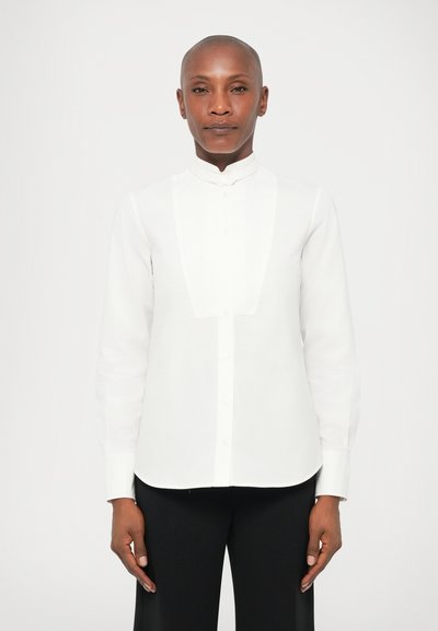 Chemise blanche à manches longues avec un col mandarin, dotée d'une patte de boutonnage sur le devant et d'un tissu lisse et léger. Associée à un pantalon noir.