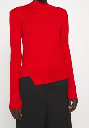 Femme portant un haut côtelé rouge vif à manches longues avec une petite fente à l'avant, associé à un pantalon noir ample.